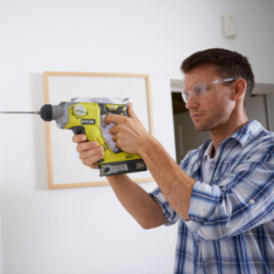 Martillo Ryobi SDS+ siendo usado por un hombre con gafas de seguridad, taladrando una pared.